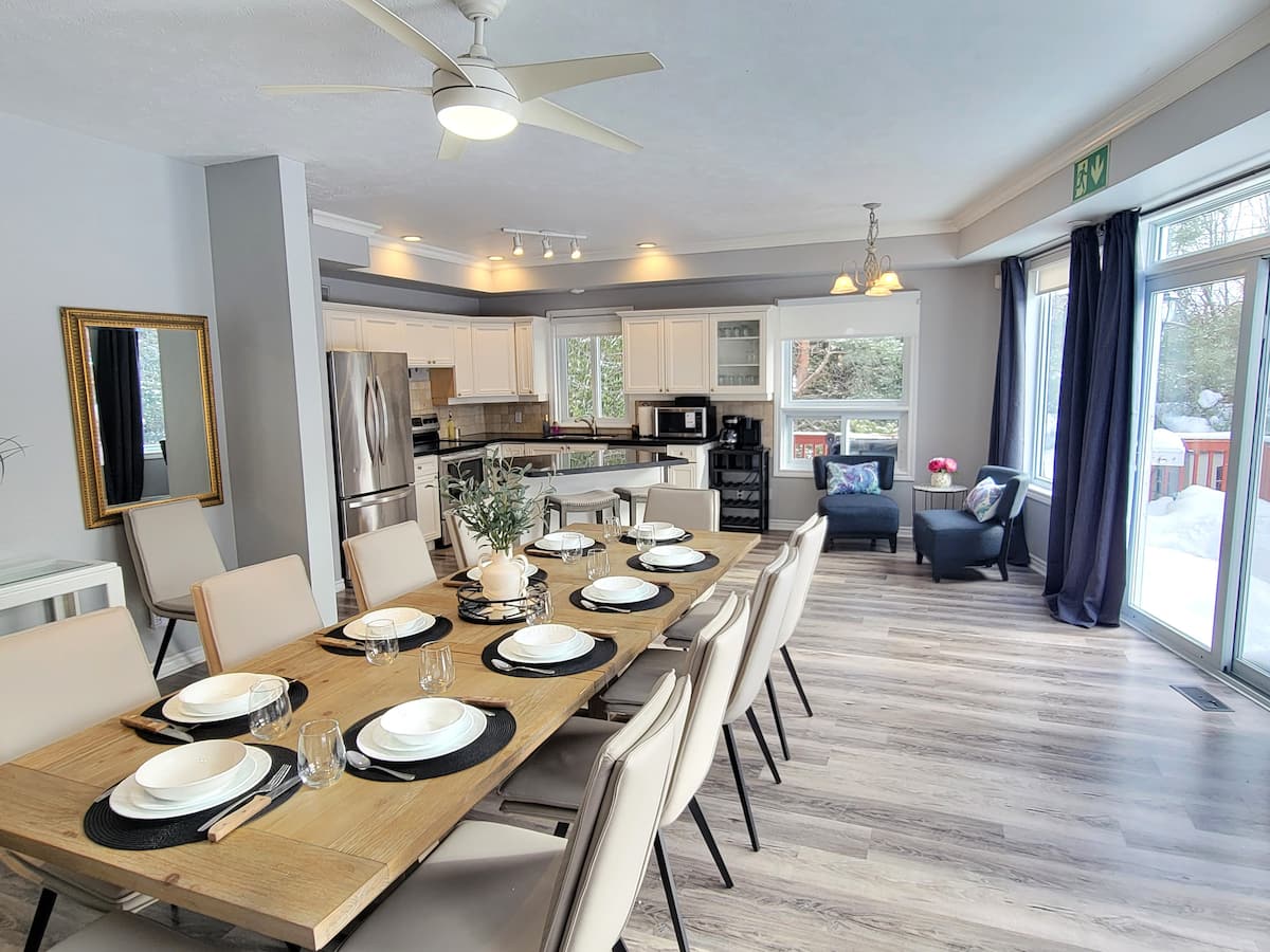 Long wooden dining table set for a group, with the kitchen behind and snow outside the sliding doors